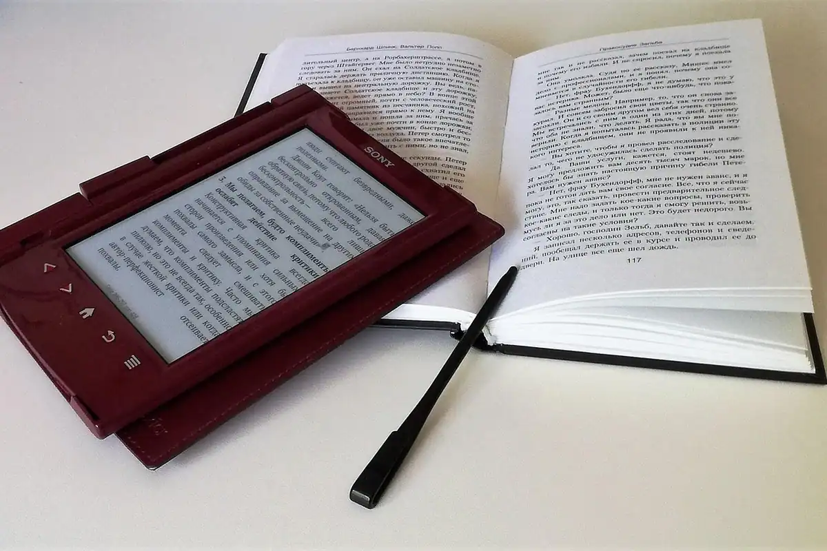 Un e-reader într-un toc roșu așezat lângă o carte tipărită deschisă, pe o masă