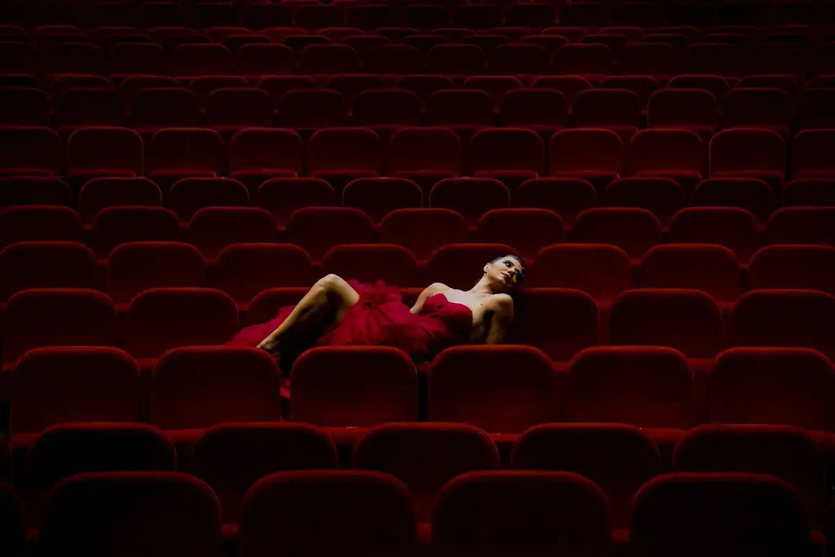 O femeie în rochie roșie stă întinsă pe scaunele de catifea ale unei săli de teatru
