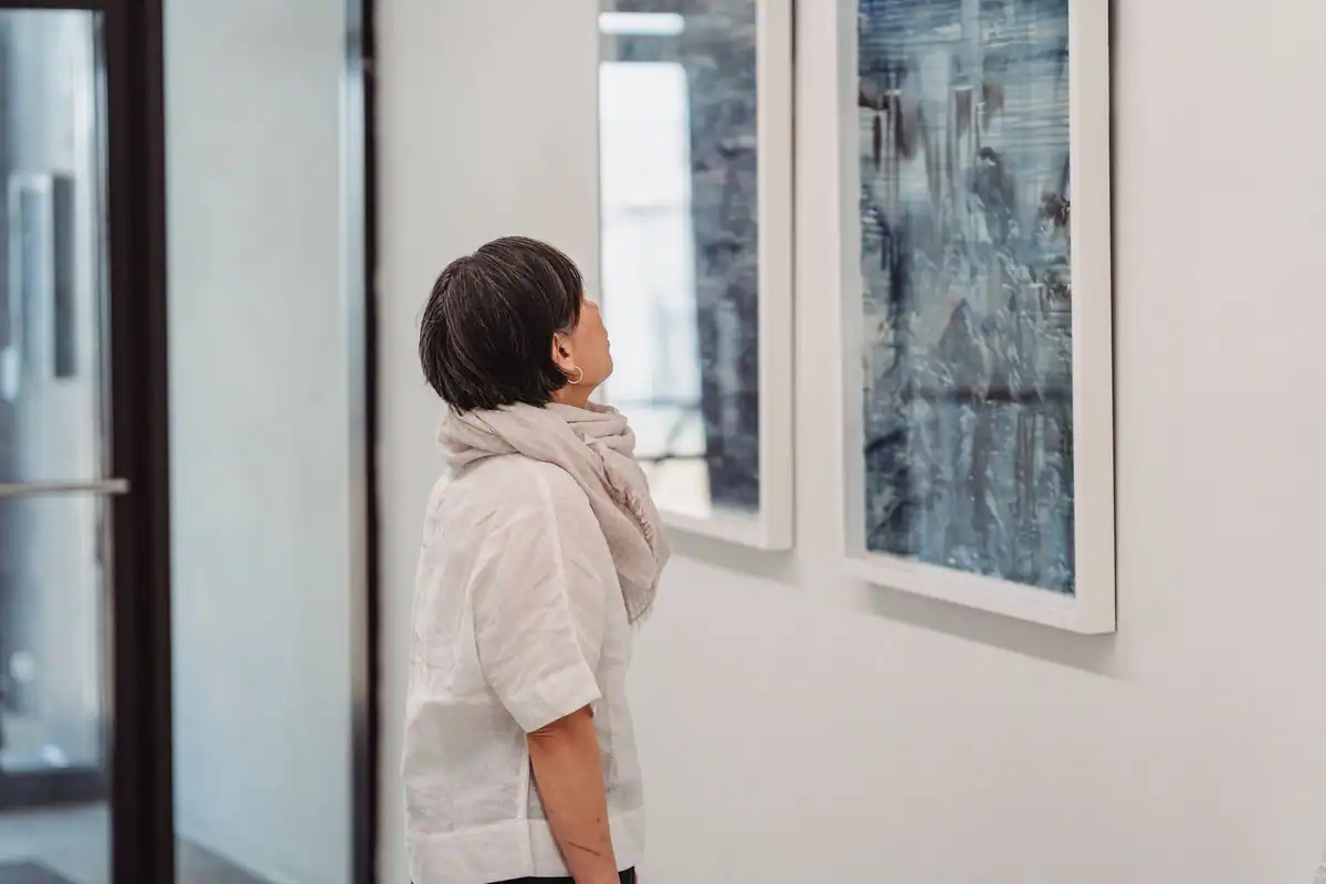 O femeie privește atentă două tablouri mari agățate pe un perete alb de galerie