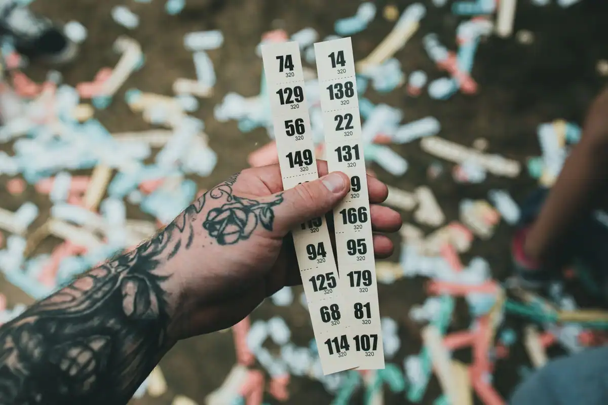 O mână cu tatuaj ține o fâșie de bilete numerotate, deasupra unor confetti colorate