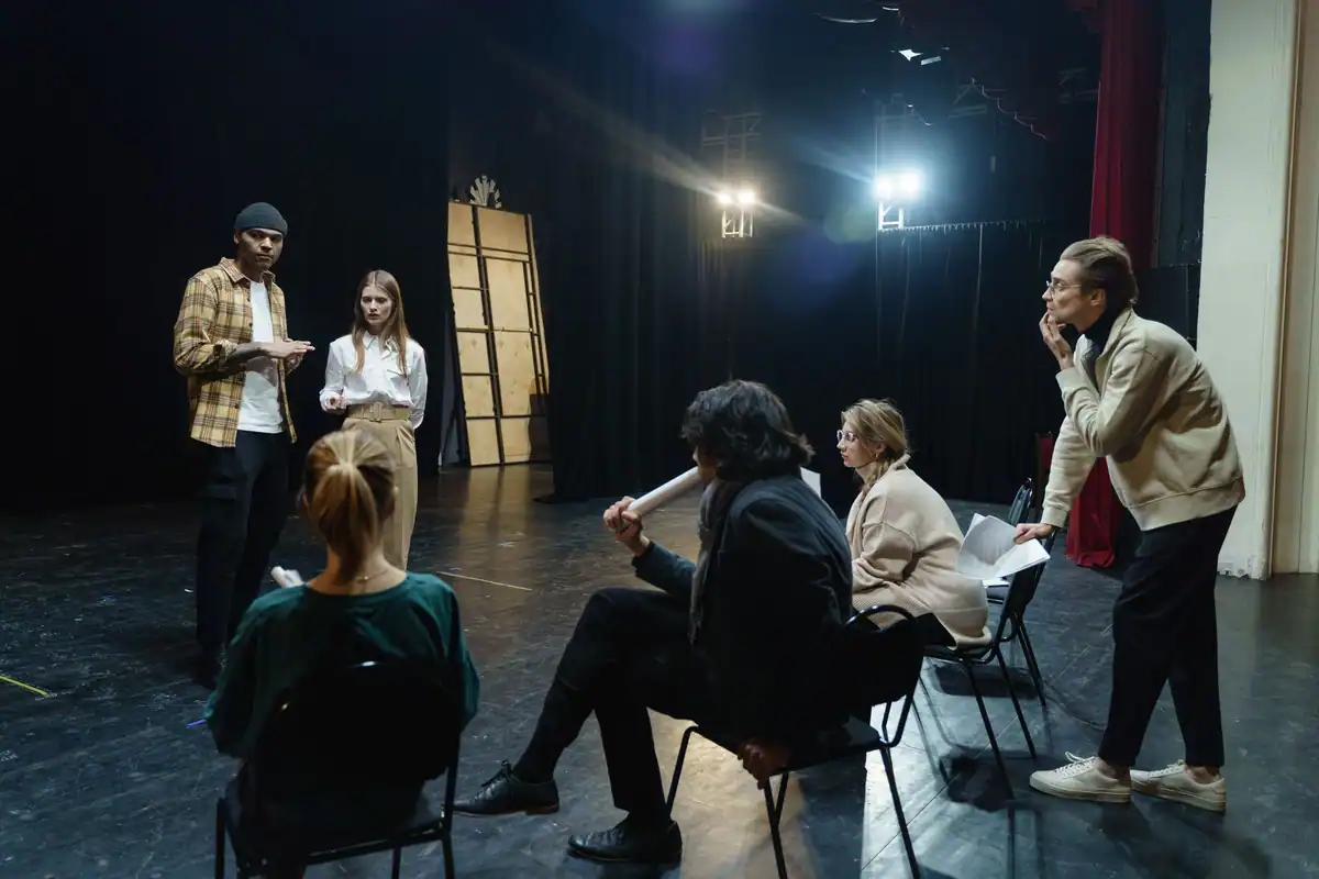 Actori discută și repetă pe o scenă de teatru, sub reflectoare, în fața scaunelor goale