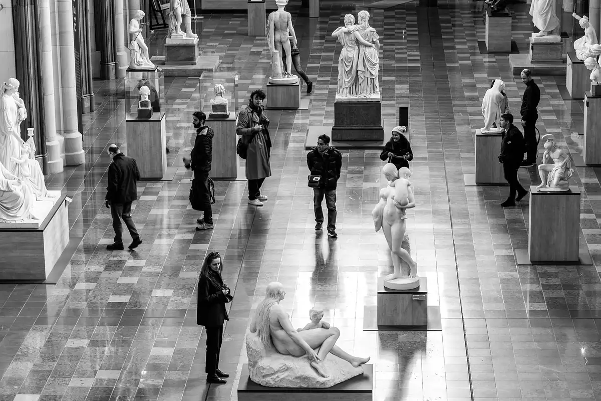 Vizitatori într-o sală largă cu statui albe pe piedestaluri, într-o fotografie alb-negru