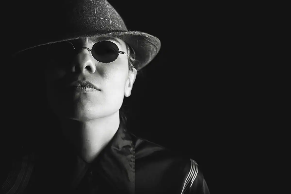 Portret alb-negru al unei femei cu pălărie și ochelari de soare, în stil film noir
