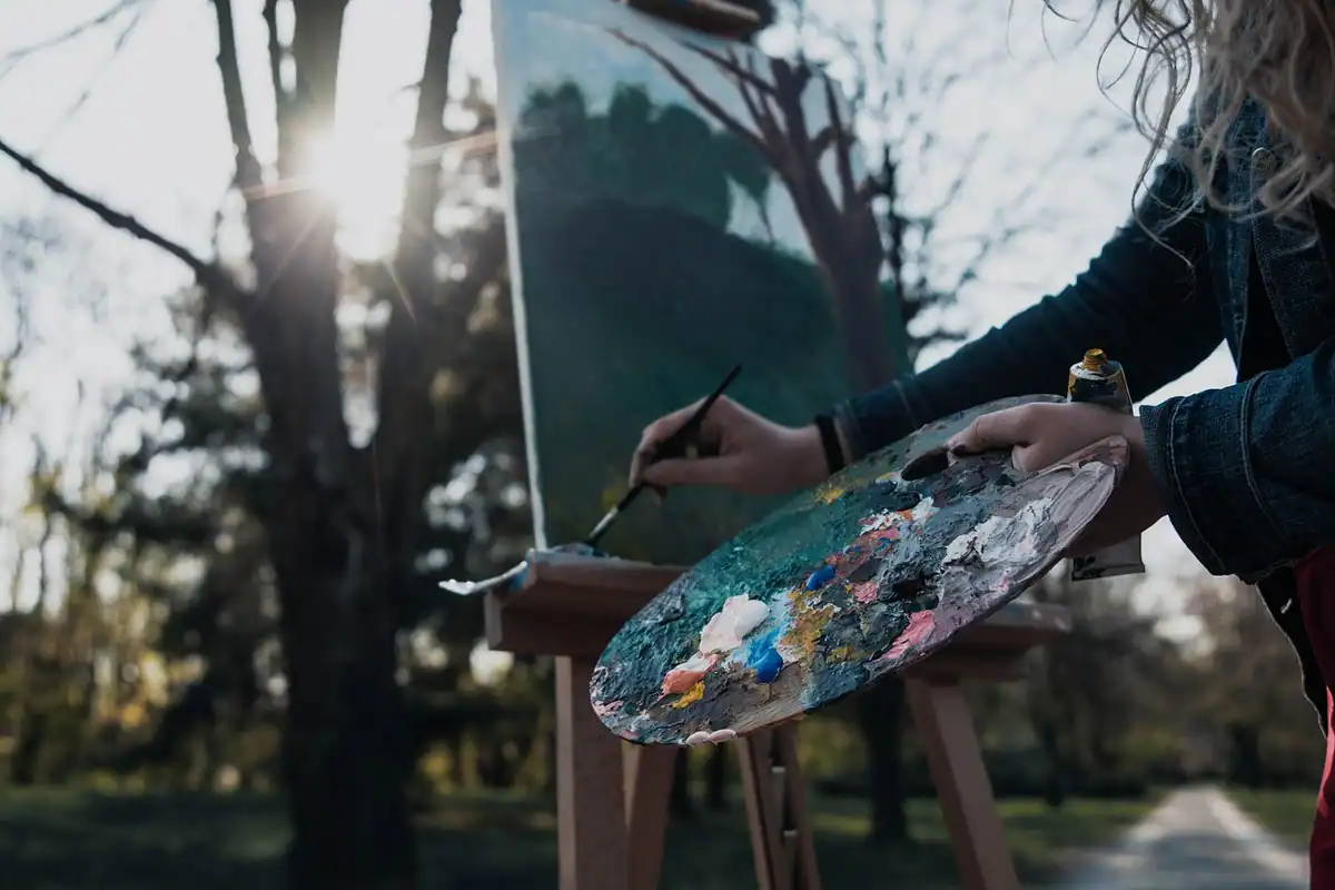 Un pictor ține paleta și pensula în fața unui șevalet, într-un parc toamna