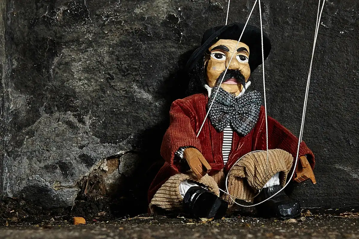 Marionetă îmbrăcată în haine roșii, atârnată de fire, lângă un zid de piatră